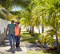 A senior couple hugging and walking down a sidewalk lined with palm trees on a sunny day.