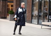 A stylish mature woman wearing a black coat and black leather boots talks on the phone outside.