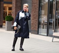 A stylish mature woman wearing a black coat and black leather boots talks on the phone outside.