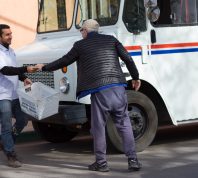 Santa Fe, NM: A senior man hands a letter to a smiling postal worker on a street in downtown Santa Fe. Close-up shot with a mail truck also in the frame.