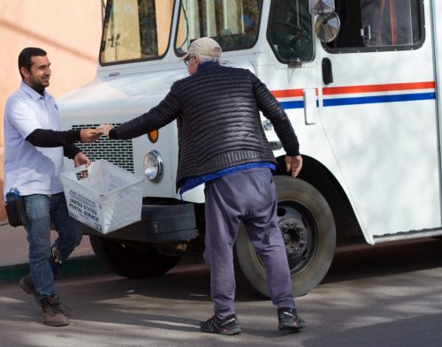 Santa Fe, NM: A senior man hands a letter to a smiling postal worker on a street in downtown Santa Fe. Close-up shot with a mail truck also in the frame.