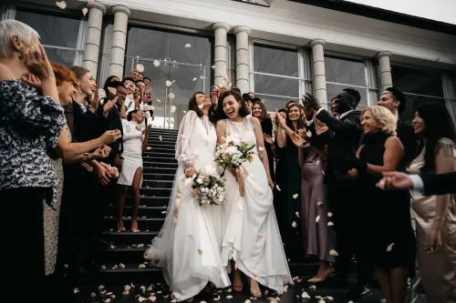 Two brides walking down steps, being showered with rose petals by wedding guests