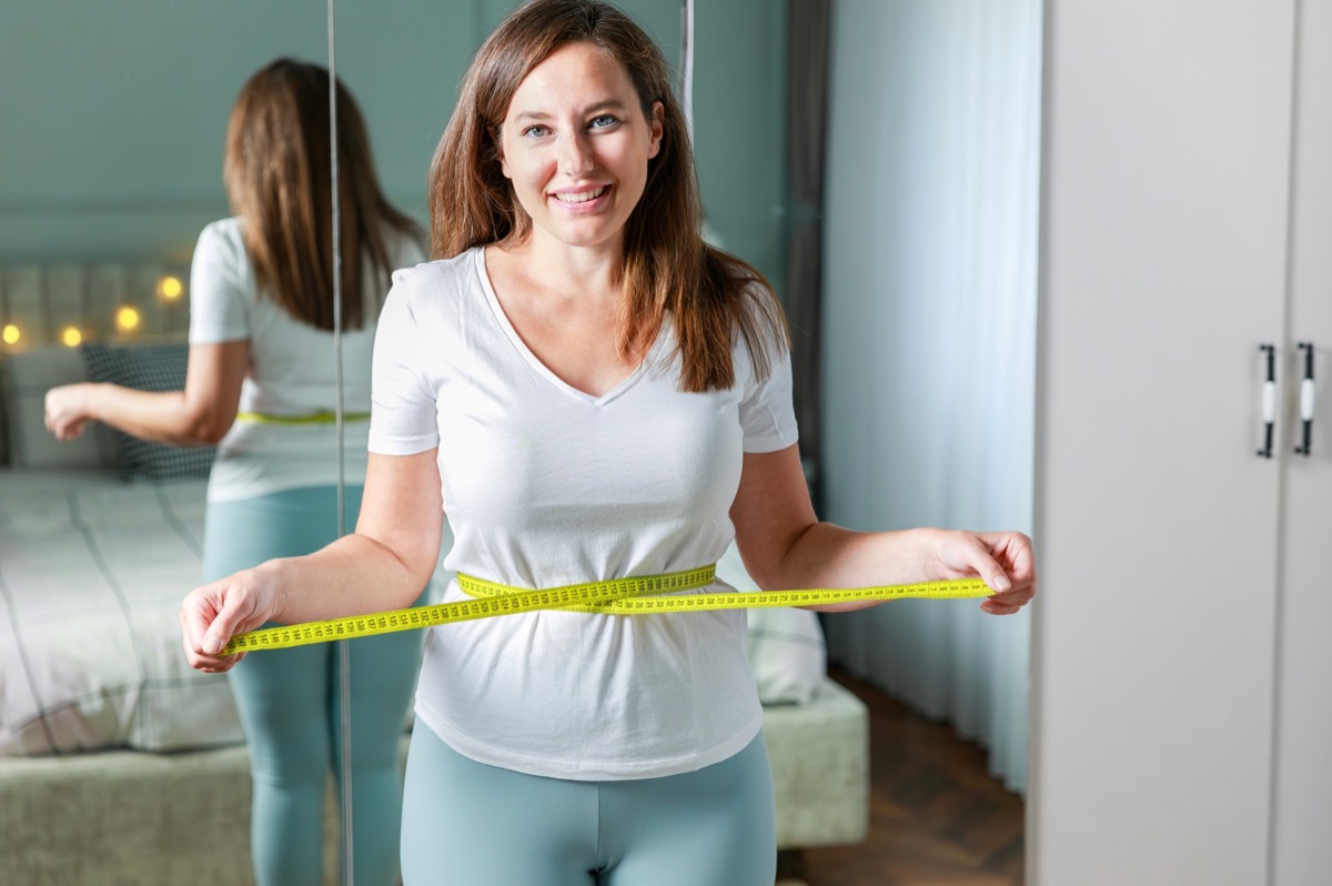 Smiling young woman after weight loss measuring waist in front of mirror