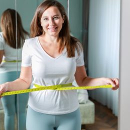 Smiling young woman after weight loss measuring waist in front of mirror