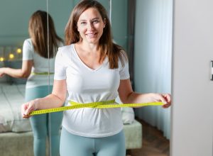 Smiling young woman after weight loss measuring waist in front of mirror