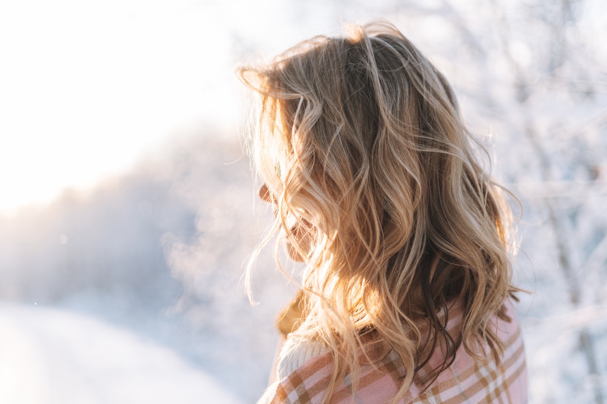 Young woman with blonde hair in winter clothes hands near face against background of snowy winter forest, view from back