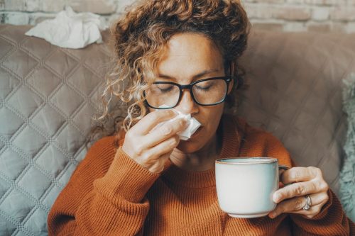 Sick woman at home blowing nose and take care of influenza virus disease. One female people using paper tissue and drink herbal tea medicine alone at home. Concept of flu cold in winter season indoor