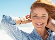 Portrait of beautiful mature woman with wind fluttering hair. Closeup face of healthy young woman with freckles looking at camera. Lady with red hair standing at seaside enjoying breeze at beach.