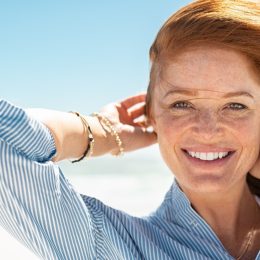 Portrait of beautiful mature woman with wind fluttering hair. Closeup face of healthy young woman with freckles looking at camera. Lady with red hair standing at seaside enjoying breeze at beach.