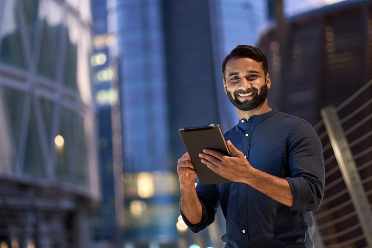 Smiling happy young eastern indian business man professional manager standing outdoor on street holding using digital tablet online fintech in night city with urban lights looking at camera, portrait.