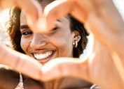 Close up image of smiling woman in swimwear on the beach making a heart shape with hands - Pretty joyful hispanic woman laughing at camera outside - Healthy lifestyle, self love and body care concept