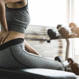 Asian Fitness woman having lower back pain after lifting too heavy dumbbells while workout. Female Athlete in grey sportswear suffering and injury from exercise at the gym. Muscle sprains.