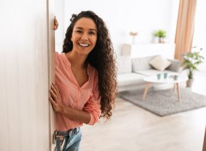 Welcome. Portrait of cheerful woman standing in doorway of modern apartment, greeting visitor and inviting guest to enter her home, happy smiling young lady holding door looking out flat