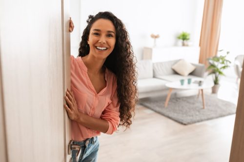 Portrait of cheerful woman standing in doorway of modern apartment, greeting visitor and inviting guest to enter her home