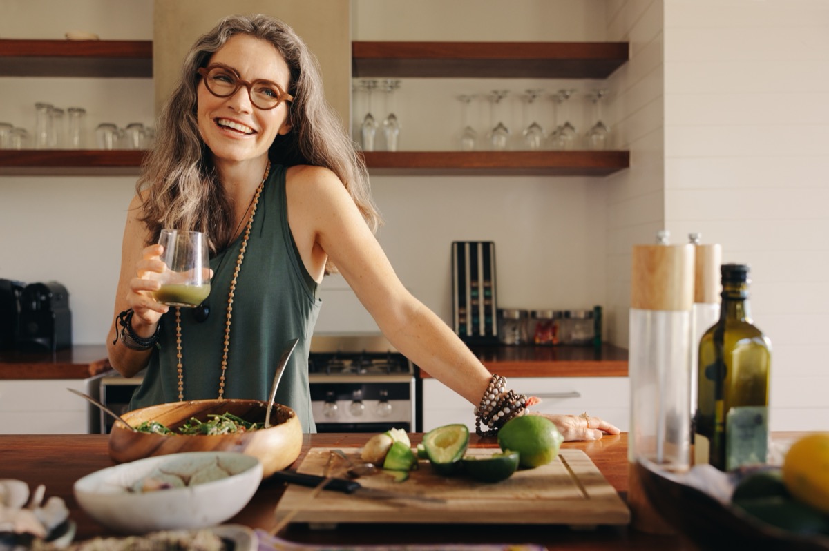 Healthy senior woman smiling while holding some green juice in her kitchen. Mature woman serving herself wholesome vegan food at home. Woman taking care of her aging body with a plant-based diet.