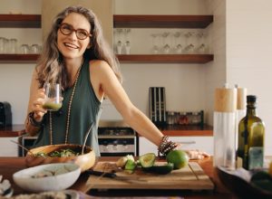 Healthy senior woman smiling while holding some green juice in her kitchen. Mature woman serving herself wholesome vegan food at home. Woman taking care of her aging body with a plant-based diet.