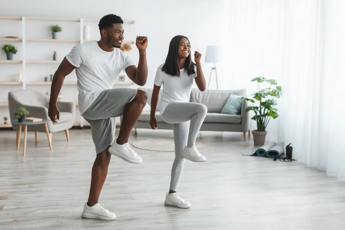 Family Workout. African American Husband And Wife Training Together In Living Room, Doing High Knees Exercise. Happy Black Couple Warming Up, Standing And Lifting Leg Up To Chest, Free Copy Space