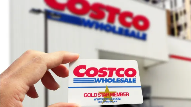 Close up of a hand holding a Costco membership card with a Costco store in the background