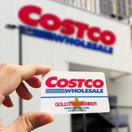 Close up of a hand holding a Costco membership card with a Costco store in the background