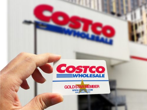 Close up of a hand holding a Costco membership card with a Costco store in the background