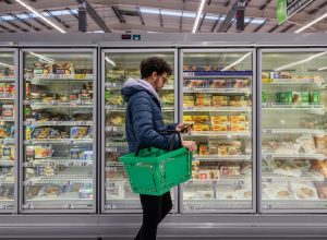 Man shopping in a supermarket while on a budget. He is looking for low prices due to inflation, standing looking at his phone in front of a row of freezers. He is living in the North East of England.