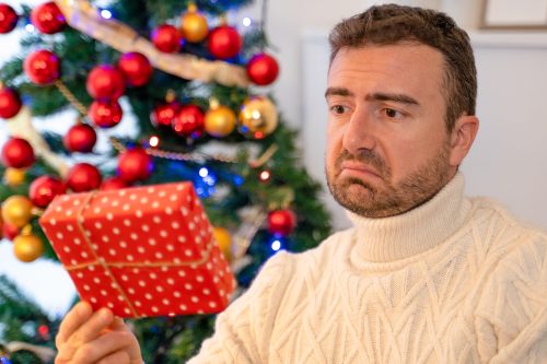 A man in front of a Christmas tree looking disappointedly at a gift.