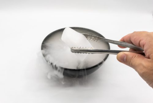 Hand holding ice tweezer with dry ice over black bowl of smoky white in motion isolated on white background.