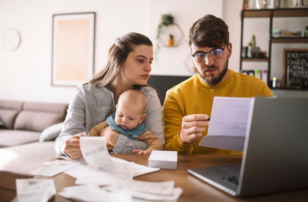 Man and woman holding a baby sitting down at the kitchen table to pay taxes or bills on their laptop