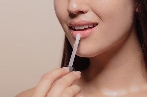 Close up of young woman applying transparent lip gloss. Cropped shot of girl putting on makeup on her lips with applicator against beige background.