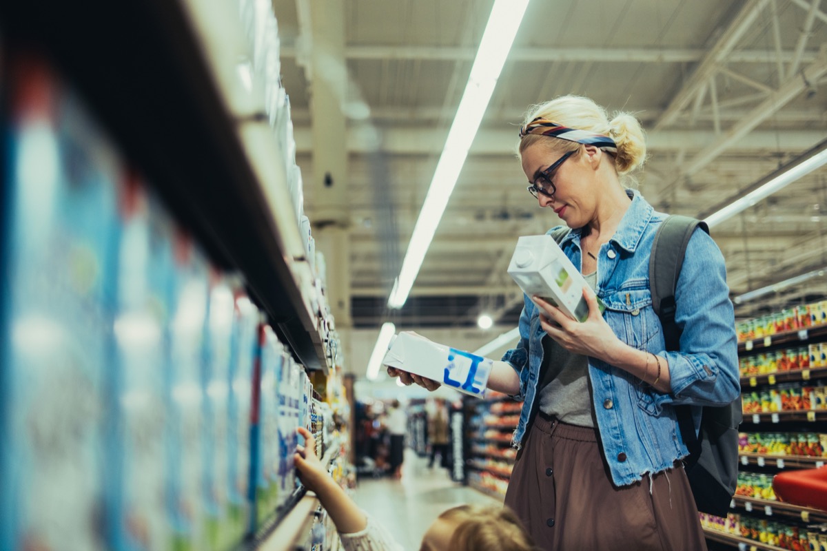 Beautiful blonde women wearing glasses, in a denim jacket and a skirt, shopping for milk in the supermarket. She is holding two cartons of milk and choosing which one to buy.