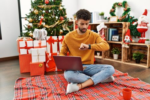 A young man sitting on the floor in front of a Christmas tree is on his laptop and checking watch, looking worried about the time.