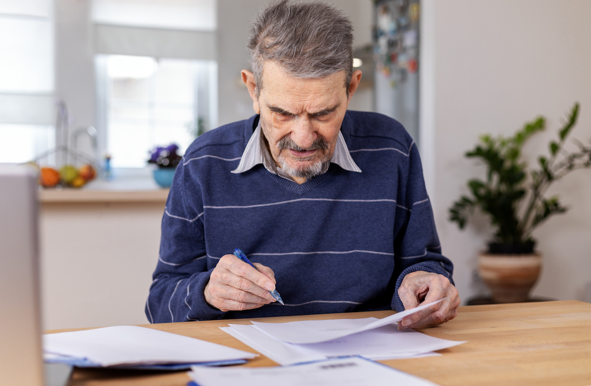 A senior adult man sitting at a table stacked with papers. He signs the documents and pays the bills