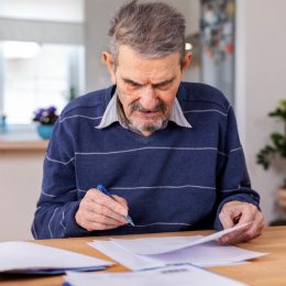 A senior adult man sitting at a table stacked with papers. He signs the documents and pays the bills