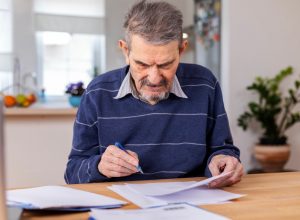 A senior adult man sitting at a table stacked with papers. He signs the documents and pays the bills