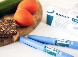 An Ozempic box and insulin injection pens next to a tray of healthy food including tomatoes, a cucumber, and bread.