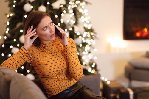 A young woman who looks perplexed is talking on her cell phone while sitting on her couch in front of the Christmas tree.