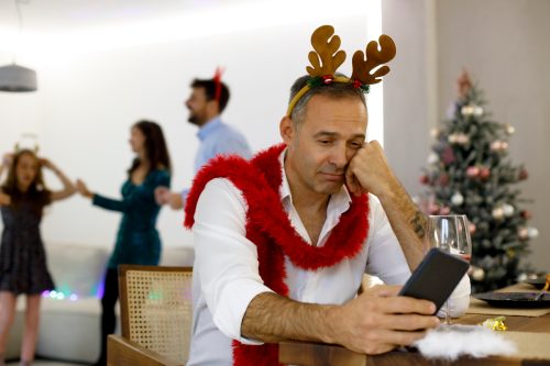 A sad-looking man wearing reindeer antlers sits along on his phone on Christmas while others celebrate in the background.