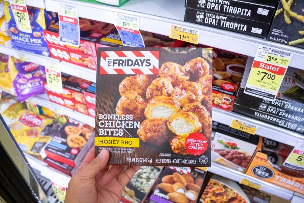 A close up of someone holding up a box of TGI Fridays Honey BBQ boneless chicken bites in a supermarket freezer section