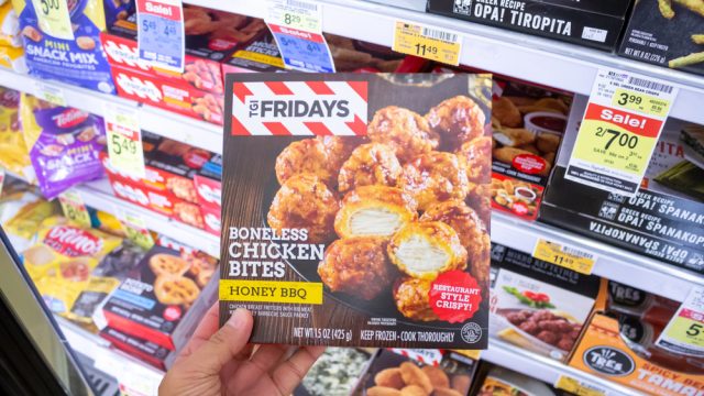 A close up of someone holding up a box of TGI Fridays Honey BBQ boneless chicken bites in a supermarket freezer section