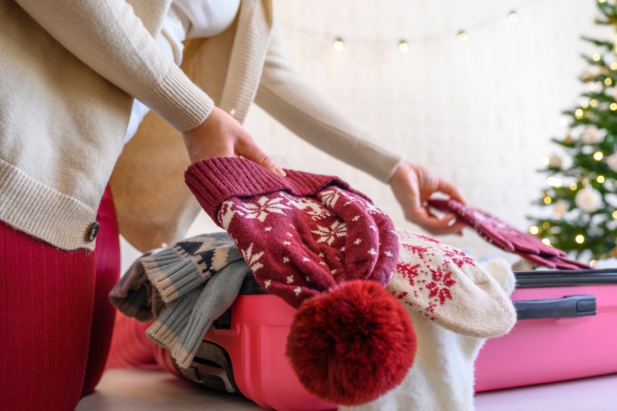 Christmas time. Woman putting knitted winter clothing in a suitcase in the room decorated christmas tree. Travel, holiday