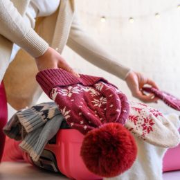 Christmas time. Woman putting knitted winter clothing in a suitcase in the room decorated christmas tree. Travel, holiday