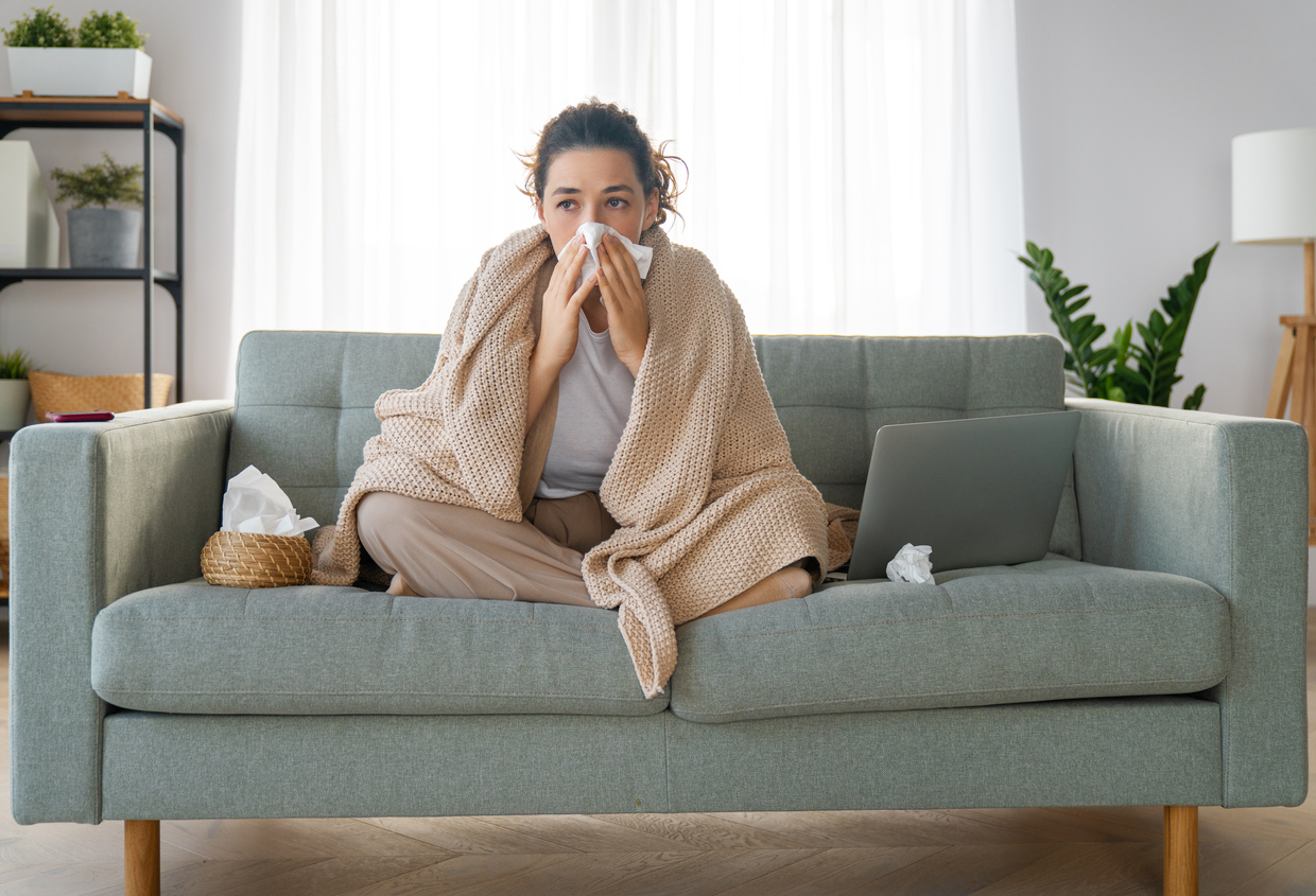 A woman sitting on the couch wrapped in a blanket while blowing her nose, probably sick