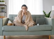 A woman sitting on the couch wrapped in a blanket while blowing her nose, probably sick