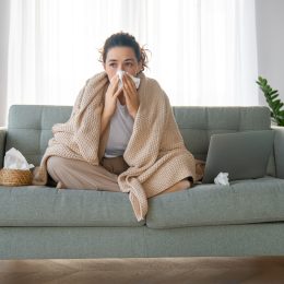 A woman sitting on the couch wrapped in a blanket while blowing her nose, probably sick