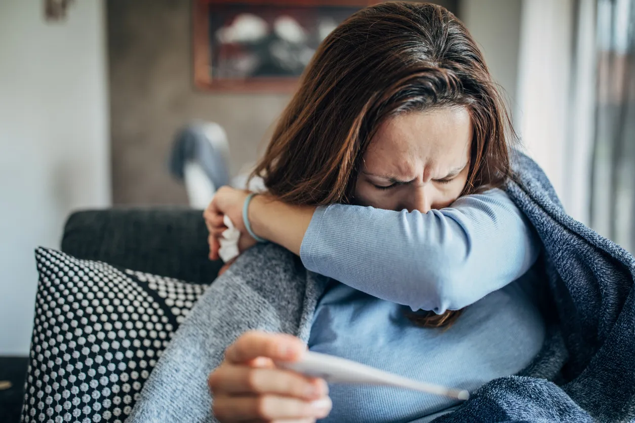 A woman coughing while holding a thermometer