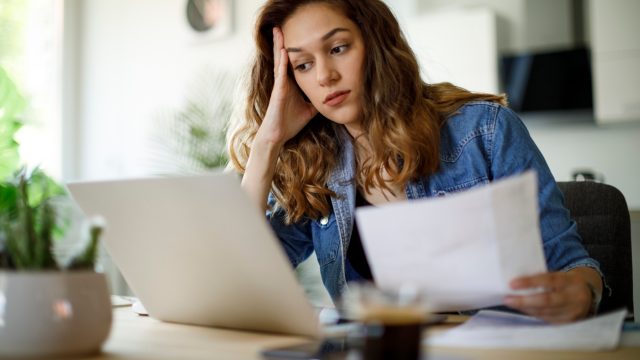 A woman using her laptop while looking over tax information or bills with a concerned look on her face