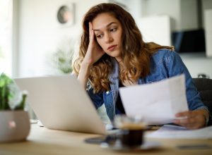 A woman using her laptop while looking over tax information or bills with a concerned look on her face