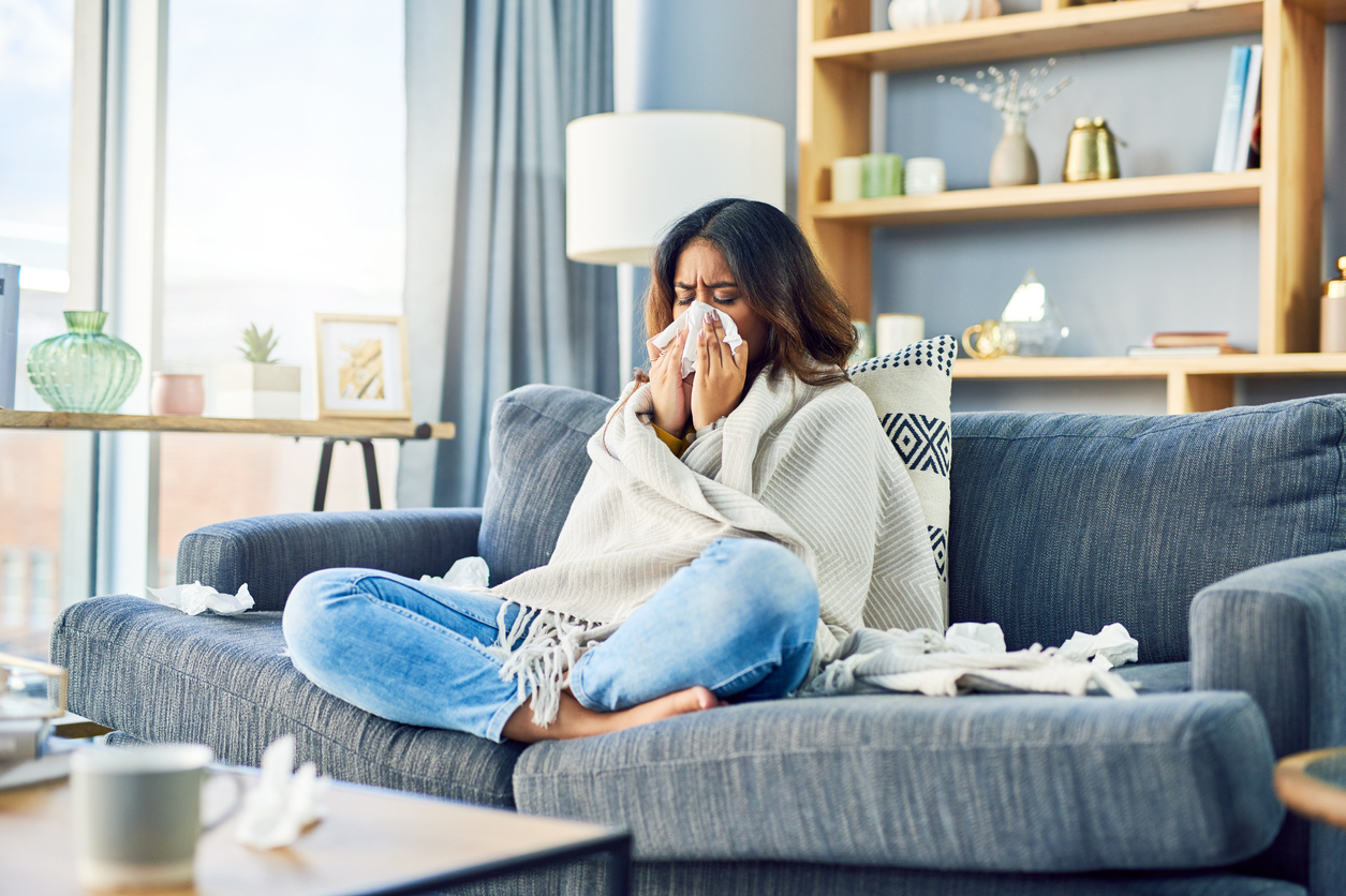 A woman sitting on the couch wrapped in a blanket and blowing her nose while surrounded by used tissues, possibly sick with the flu or COVID