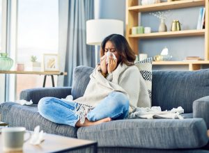 A woman sitting on the couch wrapped in a blanket and blowing her nose while surrounded by used tissues, possibly sick with the flu or COVID