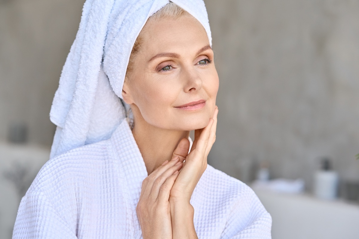 Happy beautiful middle aged woman wearing bathrobe and white towel with perfect complexion touching face looking away in bathroom. Advertising of skin care spa wellness concept. Closeup portrait.